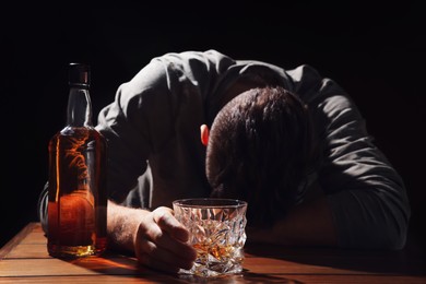 Photo of Addicted man with glass of alcoholic drink at wooden table against black background