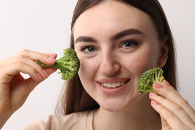Smiling woman making fake freckles with broccoli and cosmetic product on light background, closeup