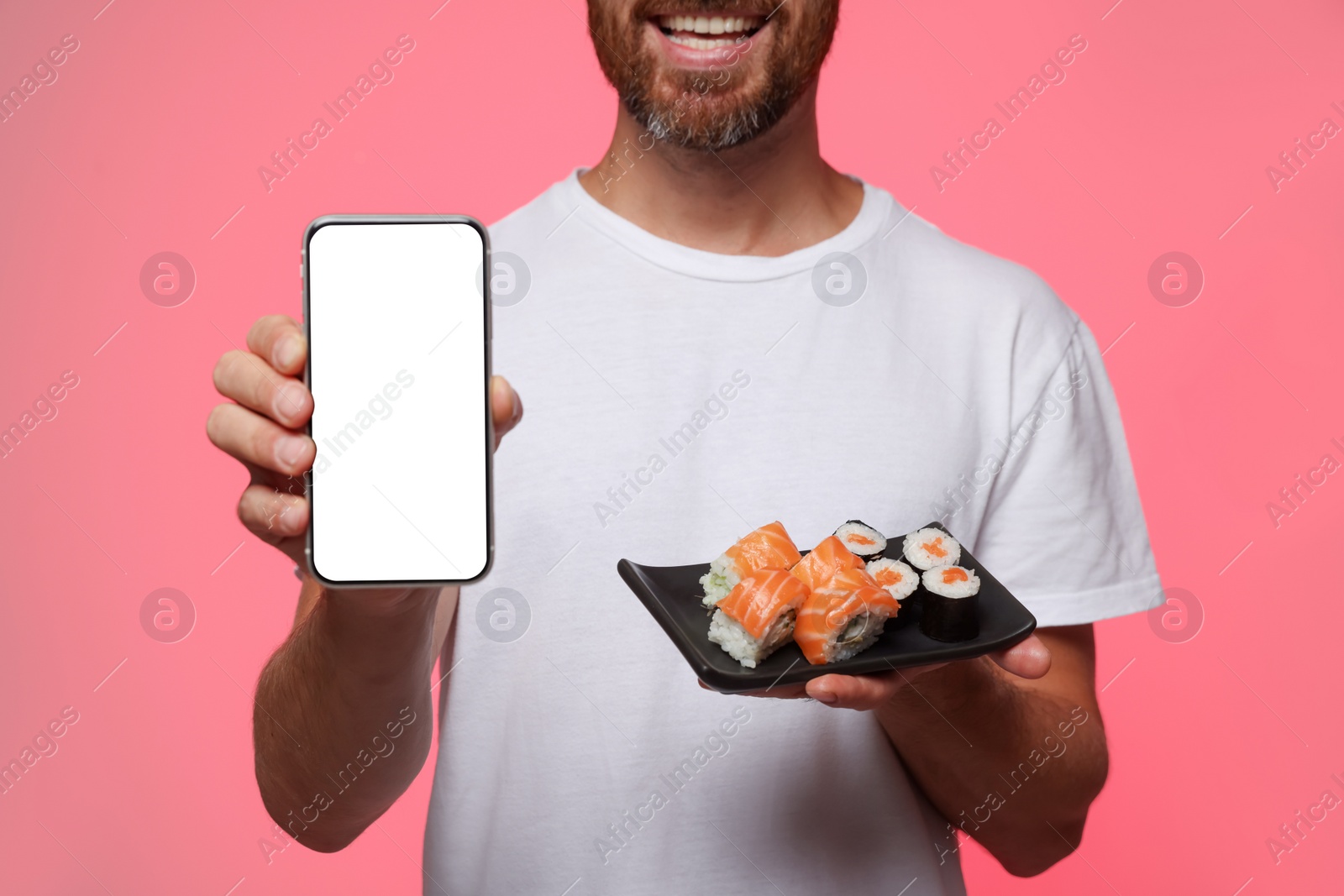 Photo of Happy man holding plate with tasty sushi rolls and smartphone against pink background, closeup