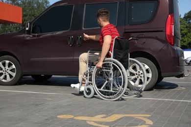 Photo of Young man in wheelchair opening door of van on car parking