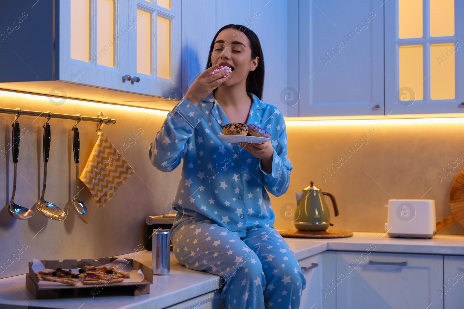 Photo of Young woman eating donut in kitchen at night. Bad habit