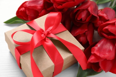 Beautiful gift box with bow and red tulip flowers on white table, closeup