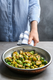 Photo of Woman with frying pan of roasted Brussels sprouts at grey table, closeup