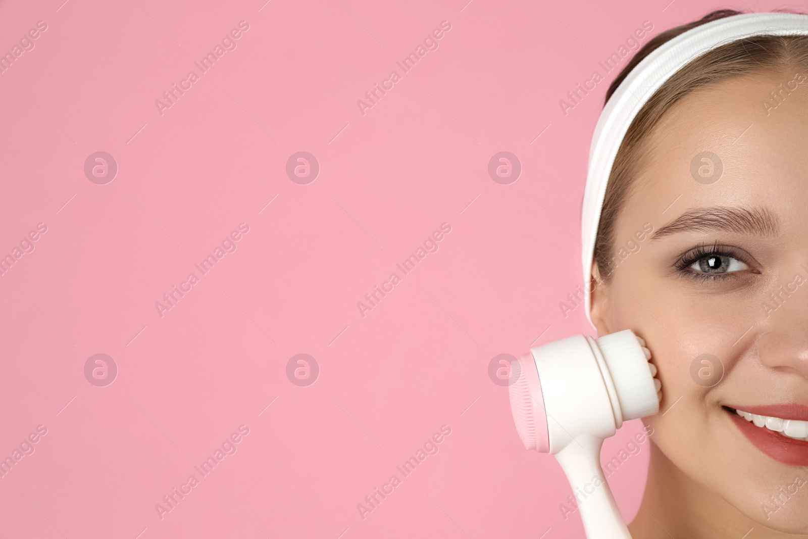 Photo of Young woman washing face with cleansing brush on pink background, space for text. Cosmetic product