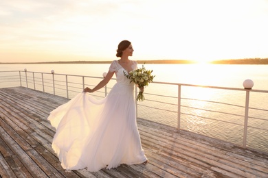 Gorgeous bride in beautiful wedding dress with bouquet near river on sunset