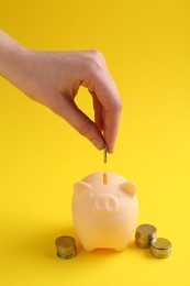 Financial savings. Woman putting coin into piggy bank on yellow background, closeup