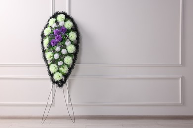 Photo of Funeral wreath of plastic flowers near white wall indoors, space for text