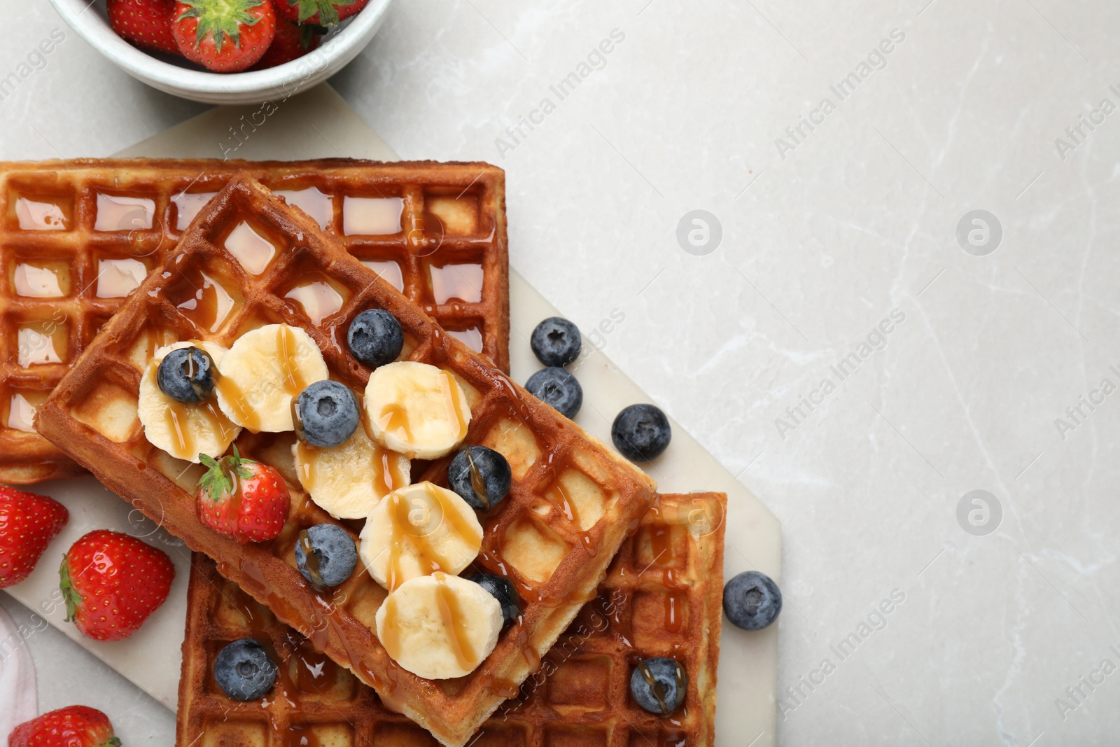 Photo of Board with delicious Belgian waffles, banana, berries and caramel sauce on light marble table, flat lay. space for text