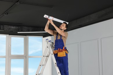 Photo of Electrician in uniform installing ceiling lamp indoors