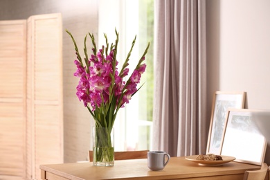 Vase with beautiful pink gladiolus flowers, pictures and cup on wooden table in room, space for text