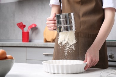 Photo of Woman sieving flour into baking dish at table in kitchen, closeup