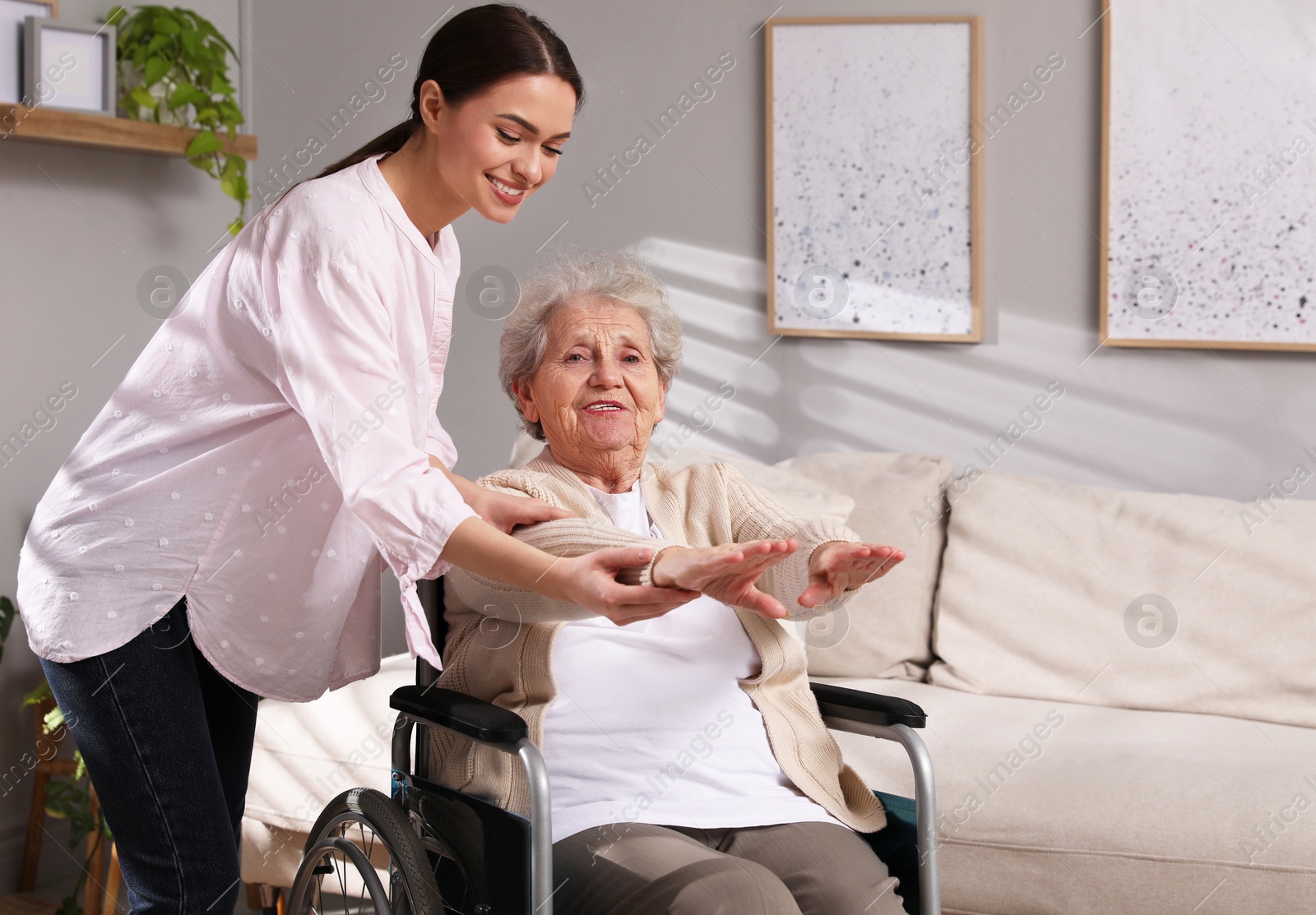 Photo of Senior woman in wheelchair doing physical exercise and young caregiver helping her indoors. Home health care service