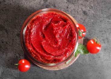 Photo of Jar of tasty tomato paste and ingredients on grey textured table, flat lay