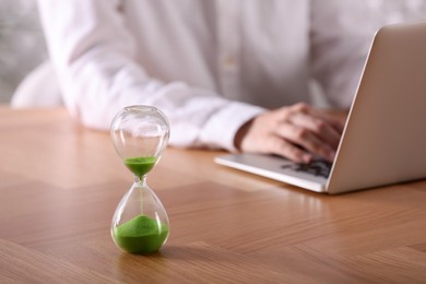 Photo of Hourglass with flowing sand on wooden table, selective focus. Man using laptop indoors