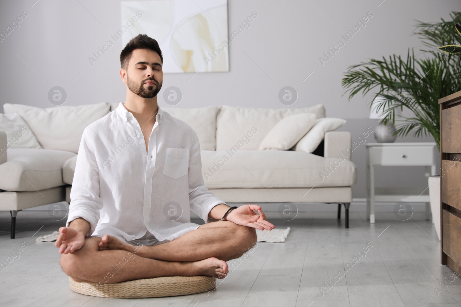 Photo of Young man meditating on straw cushion at home, space for text