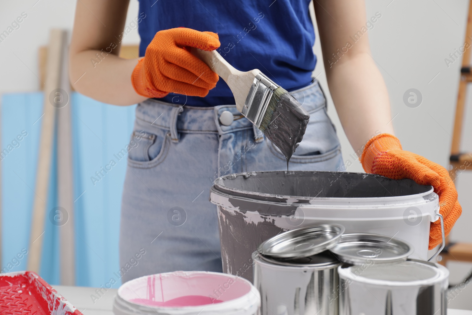 Photo of Woman dipping brush into bucket of grey paint at table indoors, closeup