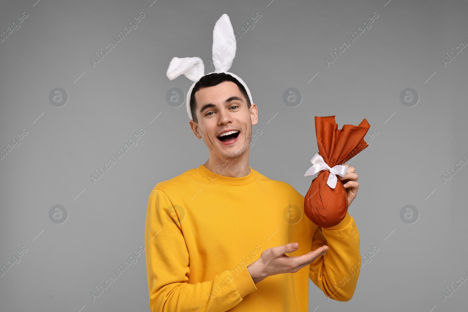 Photo of Easter celebration. Handsome young man with bunny ears holding wrapped gift on grey background