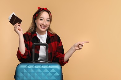 Happy young woman with passport, ticket and suitcase pointing at something on beige background, space for text