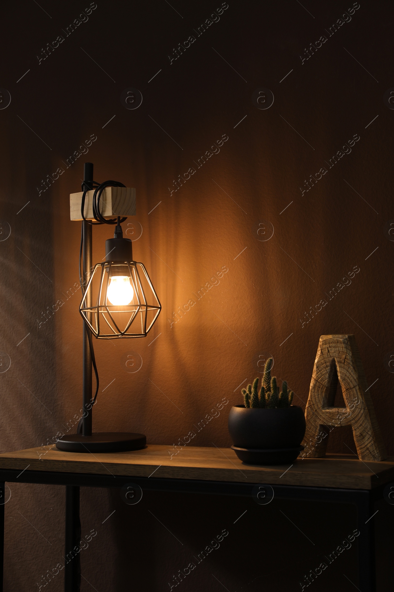 Photo of Stylish lamp, decor and green plant on wooden table near brown wall indoors. Interior design