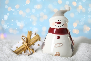 Decorative snowman near gift box on artificial snow against blurred festive lights