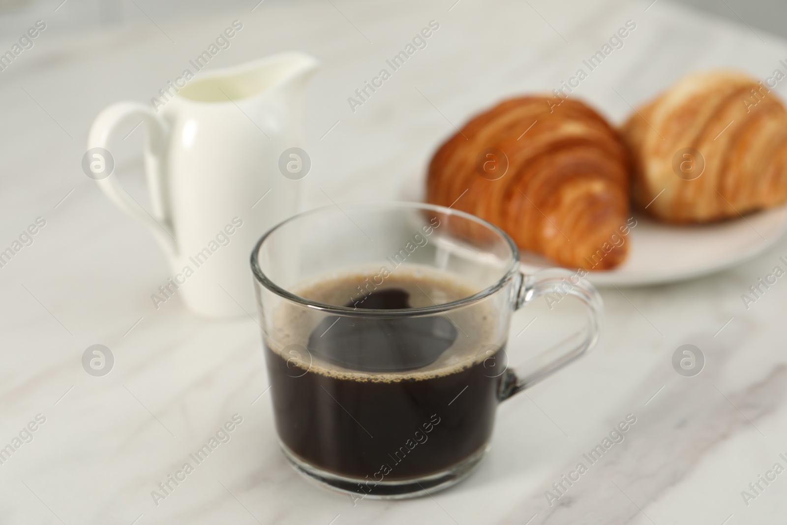 Photo of Aromatic coffee in glass cup, pitcher and fresh croissants on white marble table, closeup