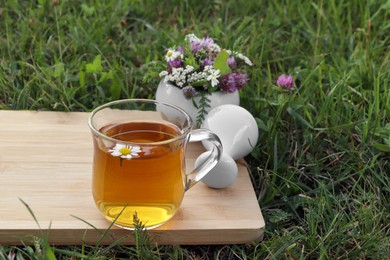 Cup of aromatic herbal tea, pestle and ceramic mortar with different wildflowers on green grass outdoors. Space for text