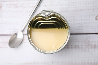 Photo of Tasty condensed milk in tin can and spoon on white wooden table, top view