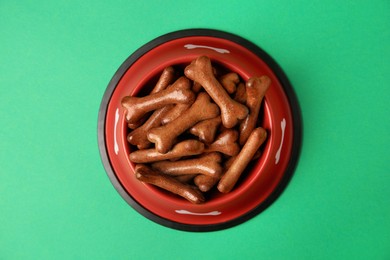 Red bowl with bone shaped dog cookies on green background, top view