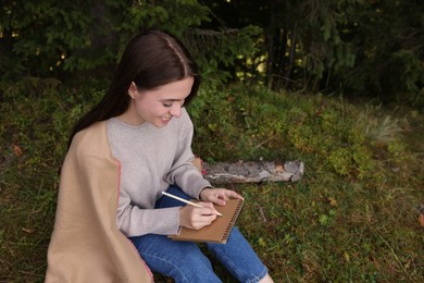 Photo of Young woman drawing with pencil in sketchbook near forest, space for text