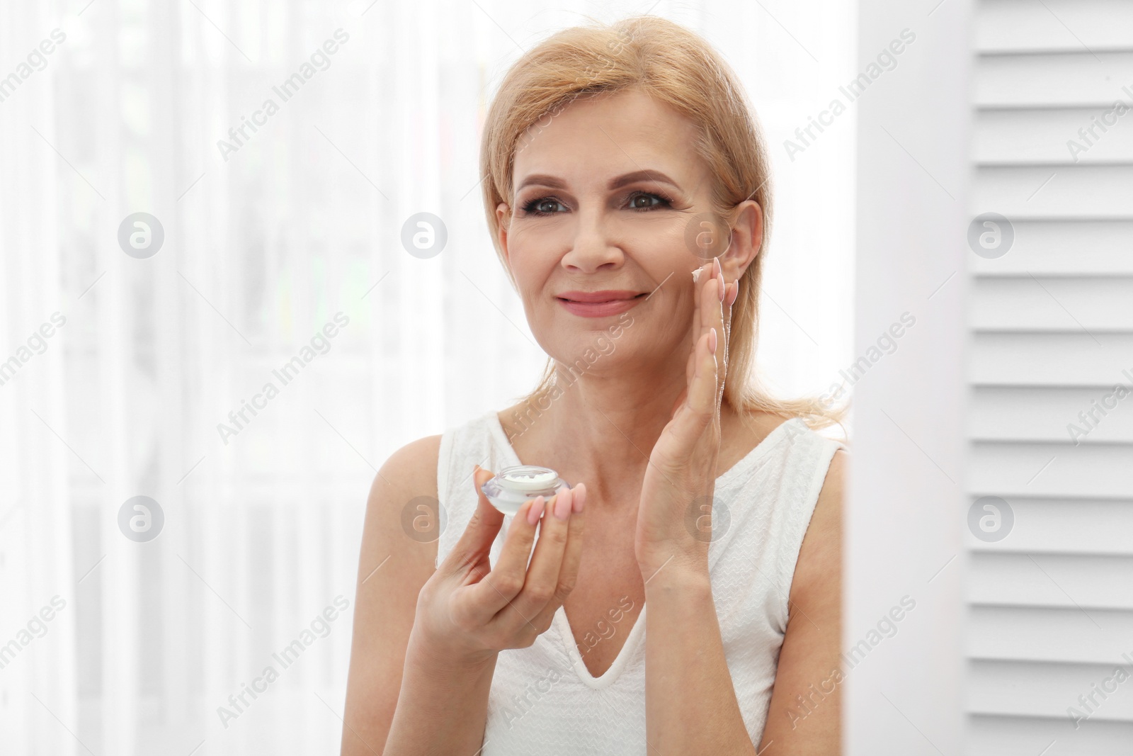 Photo of Mature woman applying face cream near mirror indoors