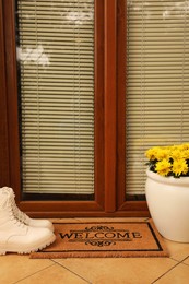 Photo of Door mat with word Welcome, stylish boots and beautiful flowers on floor near entrance