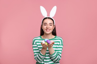 Photo of Happy woman in bunny ears headband holding painted Easter eggs on pink background