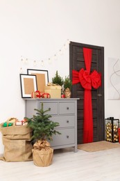 Photo of Beautiful fir tree and sack with Christmas gifts near chest of drawers and wooden door decorated with bow red indoors