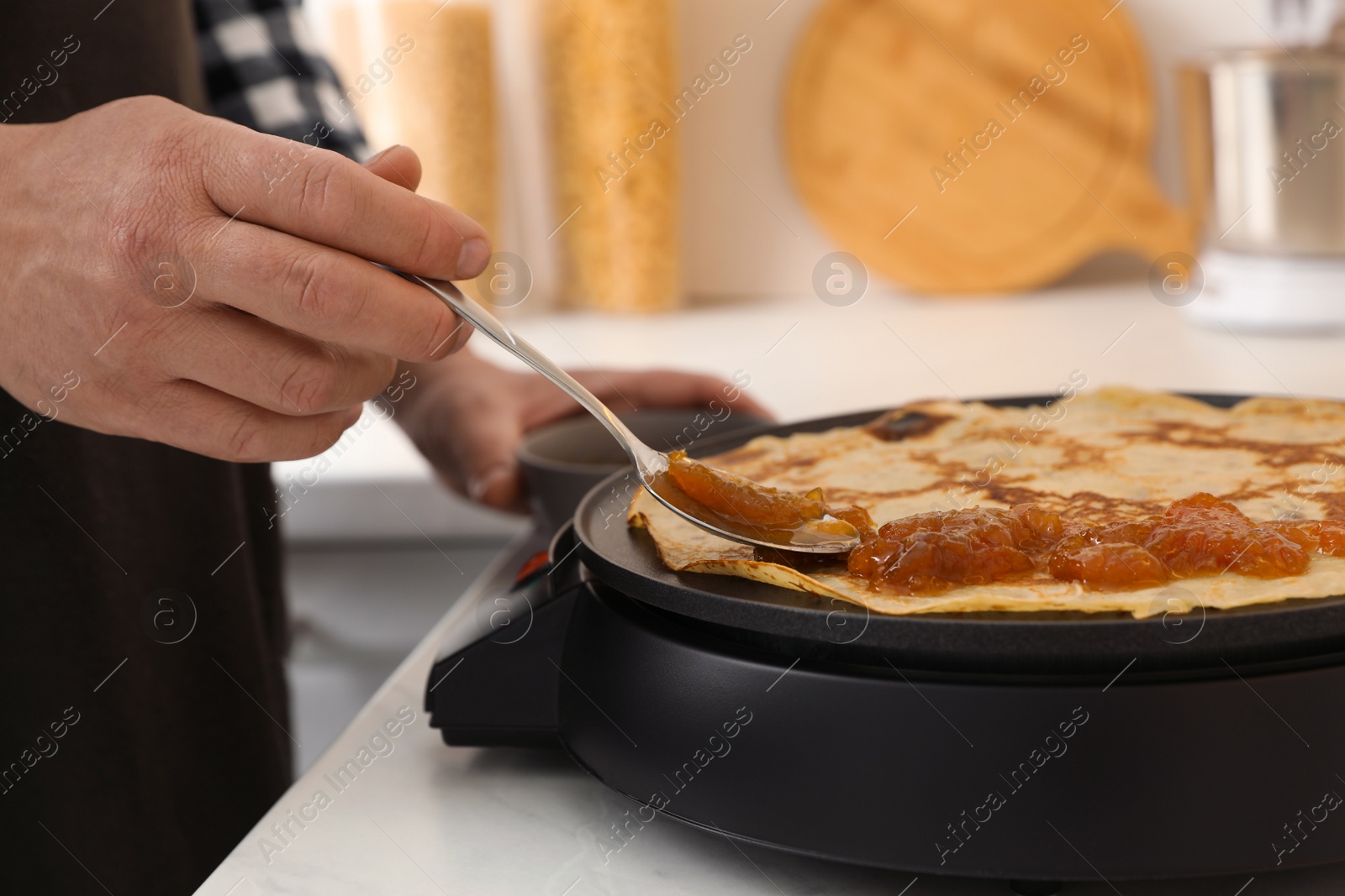 Photo of Man cooking delicious crepe with jam on electric pancake maker in kitchen, closeup