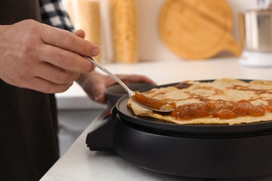 Man cooking delicious crepe with jam on electric pancake maker in kitchen, closeup