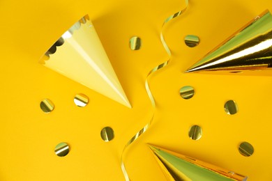 Bright party hats, serpentine streamer and confetti on yellow background, flat lay