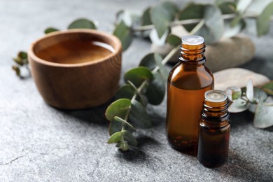 Photo of Bottles of eucalyptus essential oil, wooden bowl and plant branches on light grey table, space for text