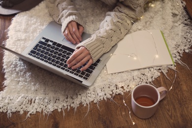 Photo of Woman with cup of hot beverage using laptop at home in winter evening, closeup