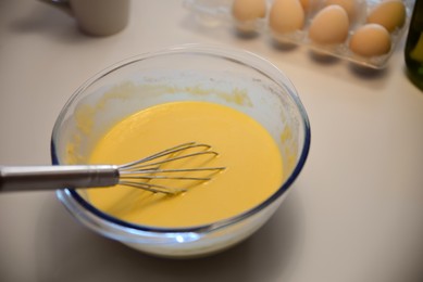 Photo of Glass bowl of crepe batter with whisk on white table, closeup