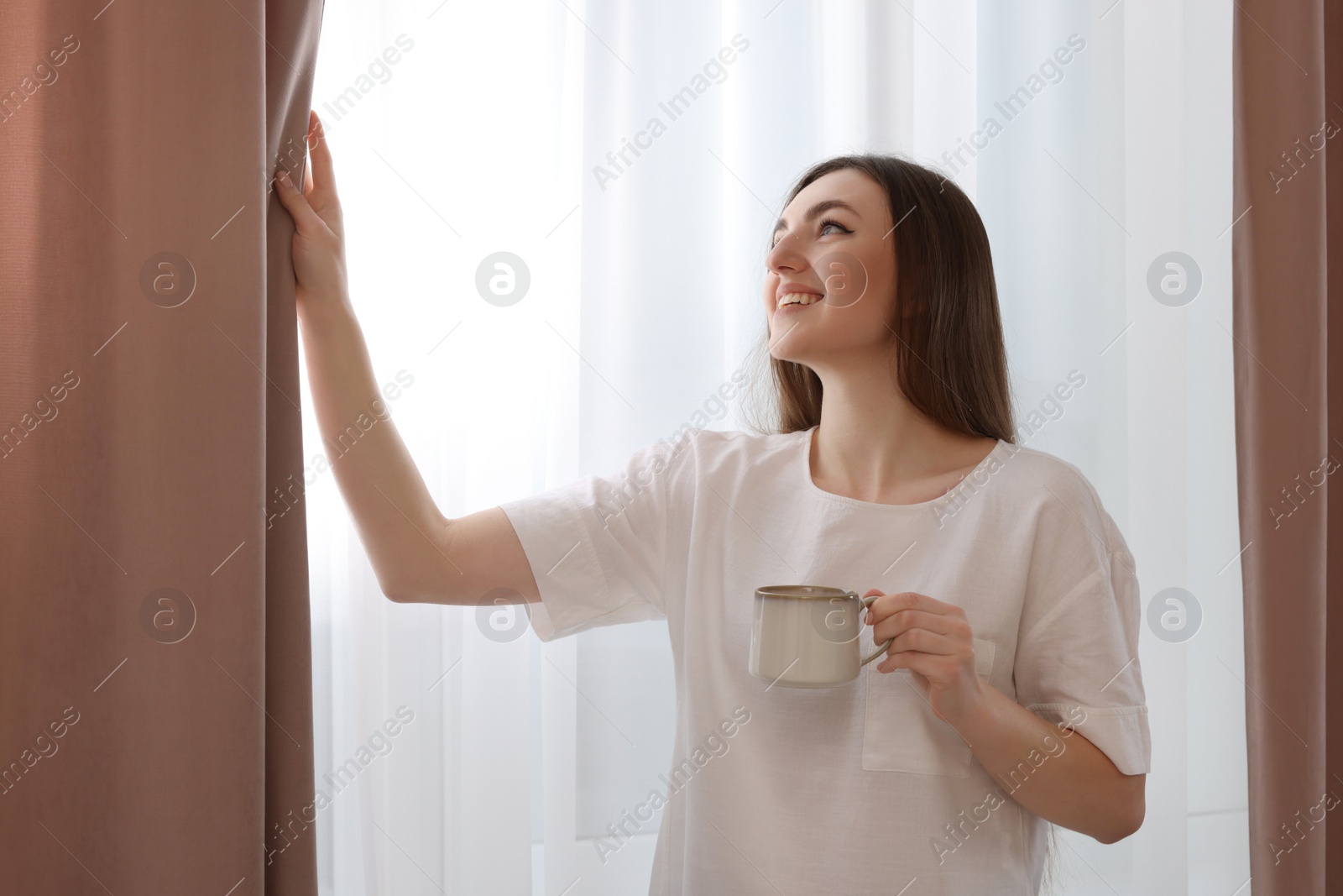 Photo of Woman with cup of hot drink opening stylish curtains at home