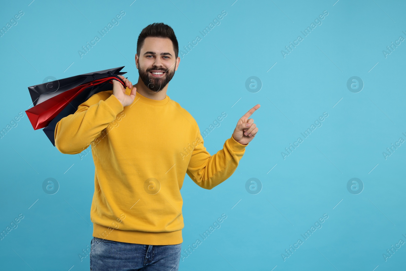 Photo of Smiling man with paper shopping bags pointing at something on light blue background. Space for text