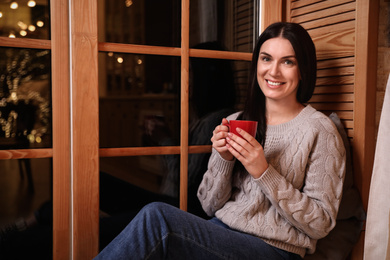 Beautiful woman with cup of coffee near window indoors. Christmas celebration
