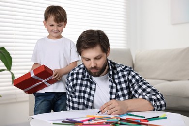 Photo of Happy Father's Day. Dad drawing at table while smiling son with gift box approaching him at home