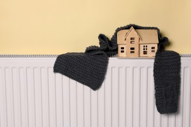 Photo of Wooden house model and knitted scarf on heating radiator near beige wall. Energy efficiency concept