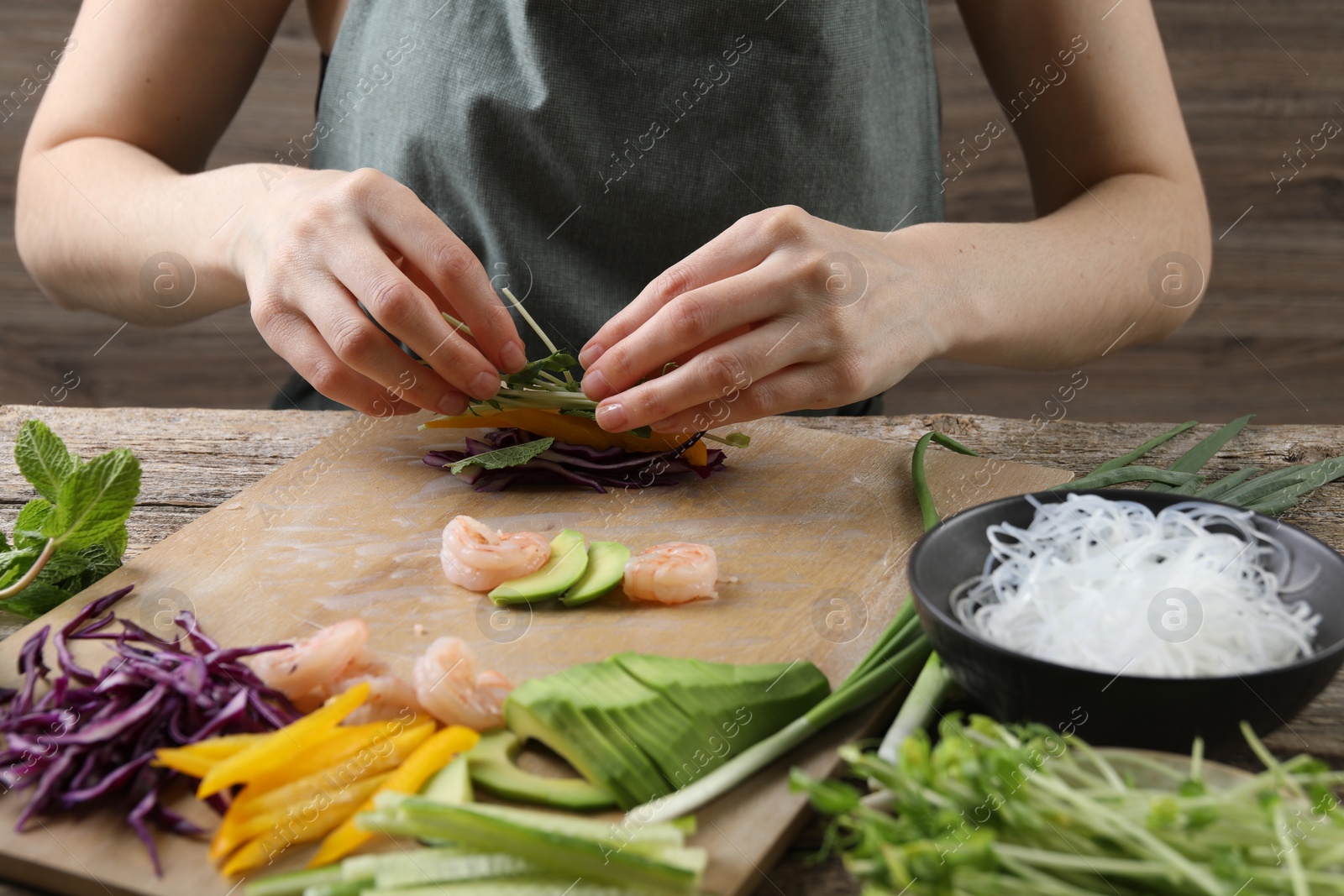 Photo of Woman wrapping spring roll at wooden table with products, closeup