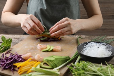 Photo of Woman wrapping spring roll at wooden table with products, closeup