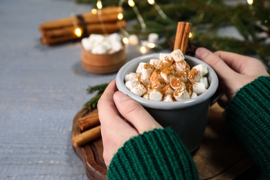Photo of Woman holding cup of hot drink with aromatic cinnamon and marshmallows at grey wooden table, closeup