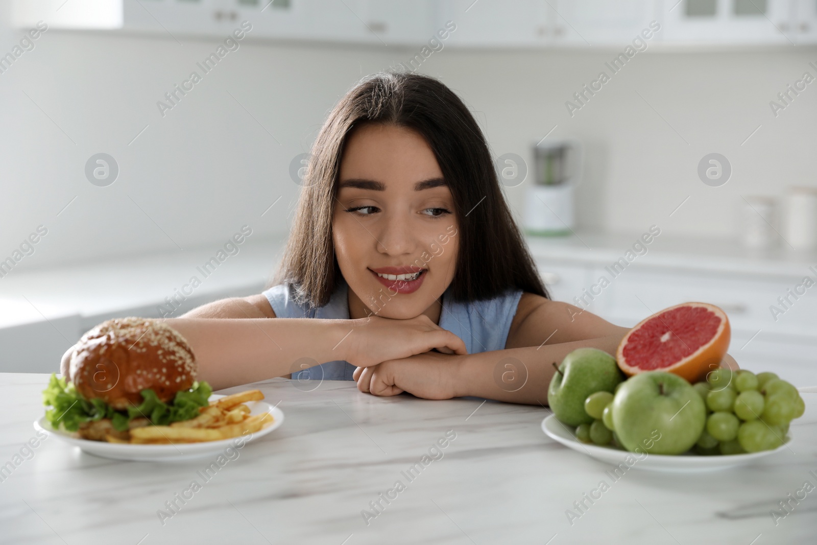 Photo of Woman choosing between fruits and burger with French fries in kitchen