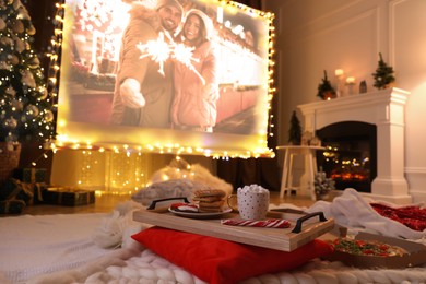 Image of Video projector screen displaying romatic Christmas movie in room, focus on tray with snack and drink. Cozy winter holidays atmosphere