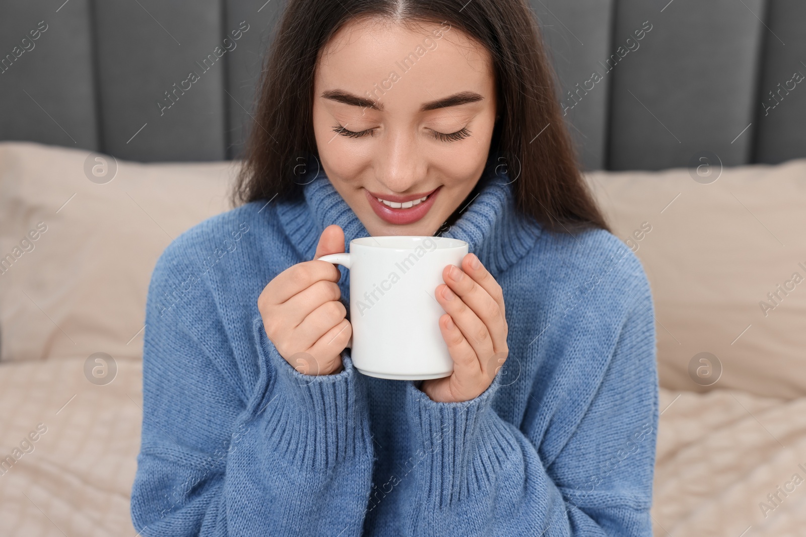 Photo of Happy young woman drinking beverage from white ceramic mug on bed